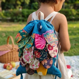 May include: A blue drawstring backpack featuring a layered petal design on the back. The petals are made from various fabrics with colorful floral and abstract patterns in shades of pink, yellow, green, and dark blue. It has white straps.