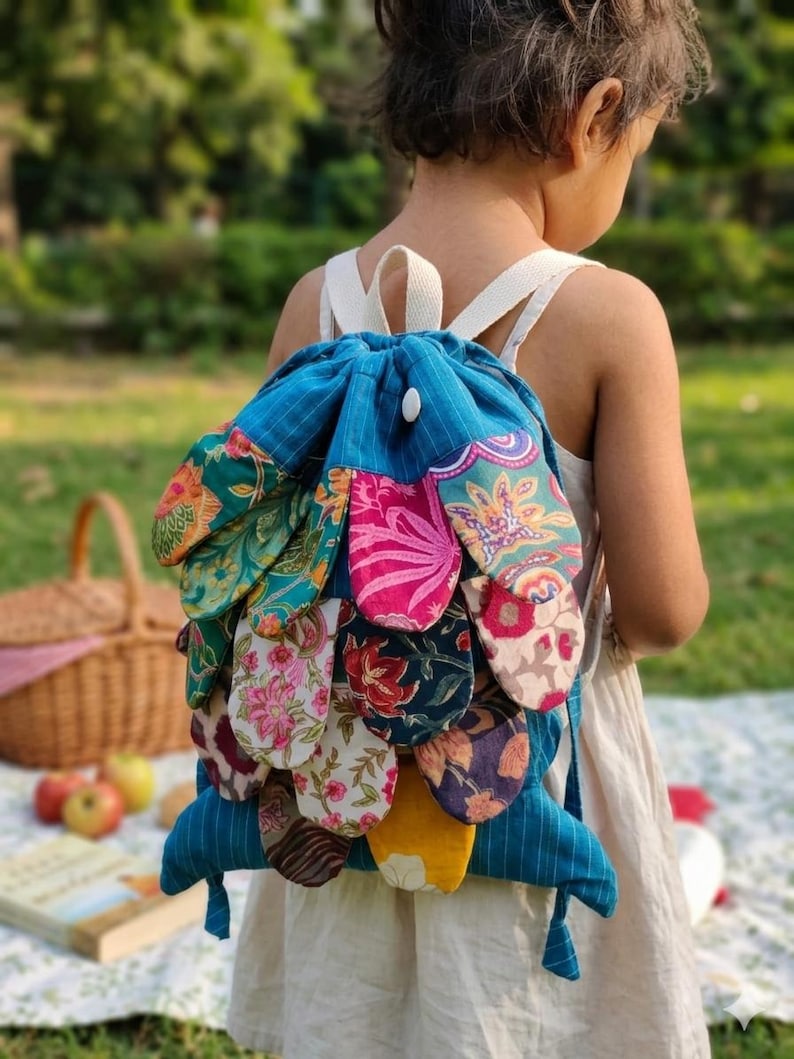 May include: A blue drawstring backpack featuring a layered petal design on the back. The petals are made from various fabrics with colorful floral and abstract patterns in shades of pink, yellow, green, and dark blue. It has white straps.