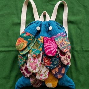 May include: A fish-shaped drawstring backpack made of blue pinstriped fabric with two white button eyes. The body is covered in overlapping fabric scales featuring various colorful floral and botanical patterns, with cream-colored shoulder straps.