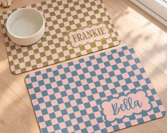 May include: Two rectangular pet mats with a checkered pattern. One mat features olive green and light pink squares with the name FRANKIE in a scalloped label. The other mat has blue and light pink squares with the name Bella in a scalloped label.