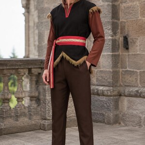 May include: A long-sleeved brown tunic worn under a black sleeveless vest with a V-neckline and brown fringe trim at the shoulders and hem. A red sash with gold embroidery is tied around the waist. The outfit includes dark brown trousers and black boots.