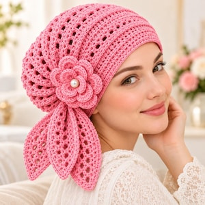 Könnte beinhalten: Eine leuchtend pinke Häkelmütze mit einem durchbrochenen Strickmuster und einer großen, mehrlagigen Häkelblume an der Seite. Die Blume ist in der Mitte mit einer weißen Perle verziert und hat zwei lange, blattförmige Häkelanhänger.