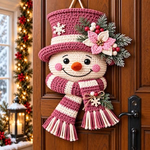 May include: A crocheted snowman wall hanging featuring a cream-colored face, a dark pink top hat with a white snowflake, and a matching striped scarf with fringe. The snowman is decorated with a pink poinsettia, red berries, and pine needles.