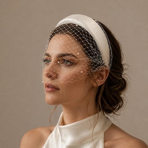 Puede incluir: Una diadema acolchada de color blanco con un velo tipo jaula incorporado. La red transparente está adornada con pequeñas perlas blancas espaciadas uniformemente por toda la malla, creando una textura delicada que cubre la parte superior del rostro.