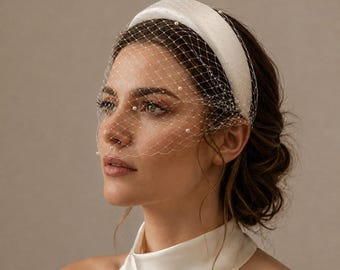 Puede incluir: Una diadema acolchada de color blanco con un velo tipo jaula incorporado. La red transparente está adornada con pequeñas perlas blancas espaciadas uniformemente por toda la malla, creando una textura delicada que cubre la parte superior del rostro.