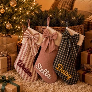 May include: Three Christmas stockings with hanging loops and bows. The first is cream with red stripes and the name Winter in red. The second is solid dusty rose with the name Stella in white. The third is black and white gingham with the name Daisy in yellow.