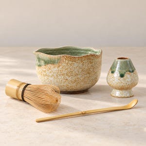 May include: A matcha tea set consisting of a ceramic bowl with a speckled beige and green glaze, a ceramic whisk holder with a matching glaze, a bamboo whisk with thin tines, and a long, slender bamboo tea scoop for measuring matcha powder.