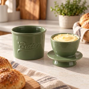 Può includere: Un burriera in ceramica verde salvia con la parola Butter in rilievo sul lato. Il set include un contenitore di base e un coperchio separato con una piccola ciotola riempita di burro giallo, progettato per mantenere il contenuto fresco.