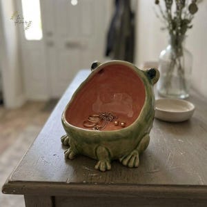 Pode incluir: Um porta-joias de cerâmica em forma de sapo com um exterior verde brilhante e um interior liso em rosa claro. A taça contém vários anéis dourados, um colar de corrente delicado e dois pequenos brincos de tarracha no seu interior.