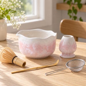 May include: A ceramic matcha tea set featuring a white and soft pink ombre bowl with a scalloped rim, a matching whisk holder, a bamboo whisk, a bamboo tea scoop, and a small stainless steel mesh strainer for preparing traditional green tea.