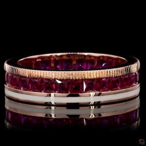 Könnte beinhalten: Ein Ring aus Roségold mit einer mittleren Reihe aus quadratisch geschliffenen, leuchtend roten Rubinen. Der Ring ist oben mit einem strukturierten, gerippten Rand aus Roségold und unten mit einem glatten, weißen Emaille-Inlay versehen.