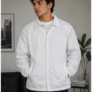 Puede incluir: Una chaqueta tipo coach de color blanco nítido con cuello clásico, mangas largas con puños elásticos y cierre frontal de botones. Esta prenda ligera presenta un acabado liso y cuenta con dos bolsillos laterales integrados.
