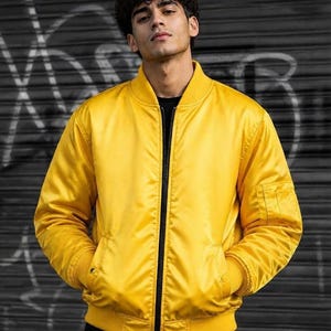 May include: A bright yellow bomber jacket featuring a black front zipper, ribbed collar, cuffs, and hem. The jacket includes two side pockets and a utility pocket on the left sleeve. The material has a smooth, satin-like finish and a solid color.