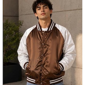 Könnte beinhalten: Eine braune Satin-Collegejacke mit weißen Raglanärmeln sowie geripptem Kragen, Bündchen und Saum mit weißen Streifen. Die Jacke verfügt über eine Druckknopfleiste auf der Vorderseite und wird über einem weißen T-Shirt getragen.