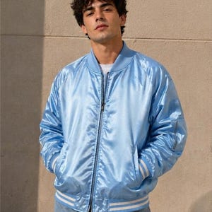 May include: A light blue satin bomber jacket featuring a front zip fastening, a ribbed collar, and ribbed cuffs with white stripes. The jacket includes long sleeves with a utility pocket on the left arm and two side pockets at the waist.