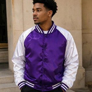 Puede incluir: Una chaqueta universitaria de satén con cuerpo de color morado intenso y mangas blancas en contraste. La prenda presenta cuello, puños y dobladillo acanalados con rayas blancas y moradas, además de un cierre frontal con botones a presión.