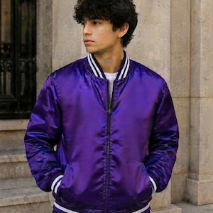 Puede incluir: Una chaqueta bomber de satén color morado vibrante con cremallera frontal completa, cuello acanalado y puños acanalados con dos rayas blancas. La chaqueta tiene un acabado suave y reflectante con un corte atlético clásico y mangas largas.