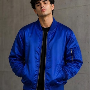 Pode incluir: Uma jaqueta bomber azul royal vibrante com gola, punhos e bainha canelados. A jaqueta possui um fecho central de zíper prateado, dois bolsos laterais e um pequeno bolso utilitário com zíper na manga esquerda, sobre uma camisa preta.
