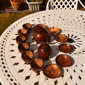 May include: A tea set crafted from dark brown coconut shells, featuring a teapot with a lid, a sugar bowl with a lid, six matching cups with handles, and six small saucers. The set displays the natural, fibrous texture of the coconut material.