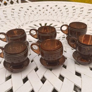 May include: A set of five dark brown coconut shell cups with matching saucers. Each cup features a rounded bowl shape, a small circular handle, and two thin horizontal engraved lines circling the exterior. The natural texture of the coconut shell is visible on the surface of each cup and saucer.