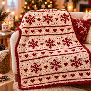 Pode incluir: Uma manta de crochê na cor creme com um padrão repetitivo de flocos de neve e pequenos corações em vermelho escuro. A manta possui uma borda grossa de crochê em vermelho escuro e está sobre um móvel, exibindo um design têxtil detalhado.