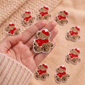 May include: A collection of small, brown, fuzzy teddy bear patches, each featuring a bright red scarf and a matching red hat. The bears have embroidered facial features and paw details, with a white border outlining the shape of each bear.