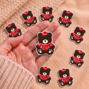 May include: A collection of small, brown bear-shaped embroidered patches. Each bear features a red cap with the word "Bear" in white, and a red and white striped shirt. The patches have a textured, fuzzy appearance and white outlines.