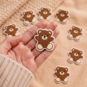 May include: A collection of small, brown, fuzzy teddy bear patches with cream-colored accents on the paws and ears. Each bear features a stitched face with a dark nose and eyes, outlined in a white border, designed for fabric application.