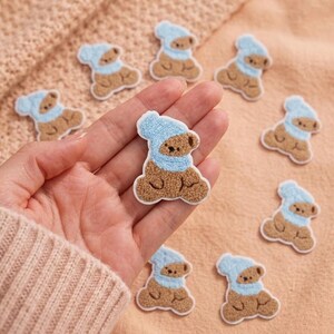 May include: A collection of small, brown, fuzzy bear-shaped patches, each wearing a light blue knitted hat and scarf. The bear patches feature embroidered details for the eyes and nose, with a textured, plush surface and a white border edge.