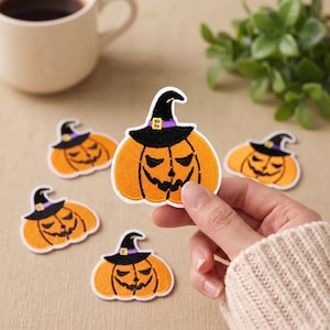 Op de afbeelding: Een geborduurde opstrijkbare patch in de vorm van een oranje pompoen met een zwart heksenhoedje, voorzien van een paarse band en een gele gesp. De patch heeft een witte rand en een gestikte textuur, met een griezelig gezicht met zwarte ogen en mond.