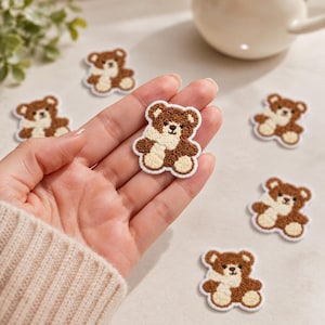 Könnte beinhalten: Eine Sammlung kleiner, braun-cremefarbener Teddybär-Aufnäher mit gestickten Details. Jeder Aufnäher zeigt einen Bären mit strukturierter Oberfläche, einem weißen Schal und einem weißen Rand. Die Aufnäher sind auf einer hellen Oberfläche angeordnet.