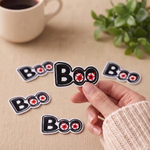 Könnte beinhalten: Ein Set aus gestickten Stoffaufnähern mit dem Wort Boo in schwarzem Garn, wobei zwei rot-weiße Augäpfel den Buchstaben o ersetzen. Die Aufnäher haben einen weißen Rand und sind zur Anbringung auf Kleidung oder anderen Textilien gedacht.