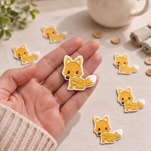 May include: A collection of small, embroidered iron-on patches featuring a stylized fox design. Each patch is rendered in shades of golden yellow and white thread with dark brown accents for the eyes and nose, showcasing a seated fox figure.