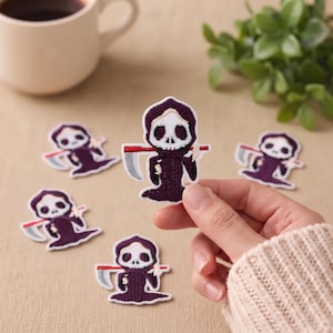 Pode incluir: Um emblema bordado apresentando um personagem de ceifador estilo desenho animado com um rosto de caveira branca, um manto com capuz roxo escuro e uma foice prateada com cabo vermelho. O emblema tem uma borda branca e é mostrado em um conjunto de cinco peças.