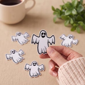 Puede incluir: Una colección de cinco parches bordados con forma de fantasma blanco, con contornos y rasgos faciales en negro. Cada fantasma tiene los brazos extendidos y un borde inferior dentado. Se sostiene un parche para mostrar los detalles.