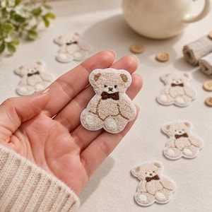 May include: A beige teddy bear patch with a textured, fuzzy surface, featuring a dark brown bow tie, small rounded ears, and white accents on the paws. The patch is shaped like a seated bear and is surrounded by several identical bear patches.