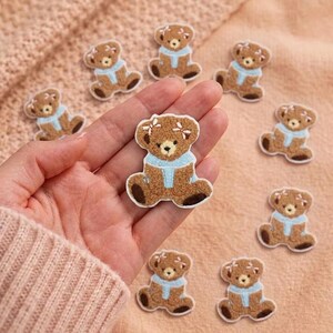 Peut inclure: Une collection de petits écussons en forme d'ours en peluche marron et duveteux, chacun portant une écharpe bleu clair et deux nœuds blancs sur la tête. Les ours sont brodés avec des pattes et des oreilles marron foncé, en appliques.