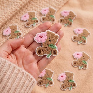 May include: A collection of small, brown, fuzzy bear-shaped patches, each featuring a textured brown bear holding a pink flower with a green stem. The patches have a white border and are arranged on a light beige fabric surface.