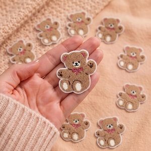 May include: A collection of small, tan, fuzzy teddy bear patches featuring embroidered details including a dark pink bow, white paw pads, and a white border. Each patch depicts a seated bear with a textured surface, designed for fabric application.