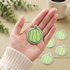 Puede incluir: Un parche bordado circular con un diseño de sandía en verde claro con rayas verticales en verde oscuro y un pequeño tallo verde en la parte superior. El parche tiene un borde blanco y se muestra en una palma, con otros parches idénticos cerca.