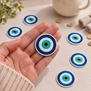 May include: A circular embroidered patch featuring a stylized eye design with a black center, a bright turquoise iris, and concentric rings of white and dark blue. The patch is round with a stitched border, showcasing a detailed thread texture.