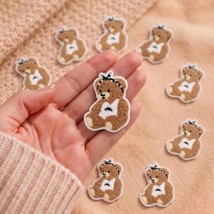 May include: A collection of small, brown, fuzzy teddy bear patches with white chest patches and black and white polka dot headbands. Each bear is depicted in a seated position with a textured, plush-like appearance and a white border outline.