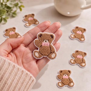 May include: A collection of small, brown, fuzzy teddy bear patches with a white border. Each bear features a textured, plush-like appearance, dark brown ears, and a light pink bow around its neck. The patches are shown individually and in a group.