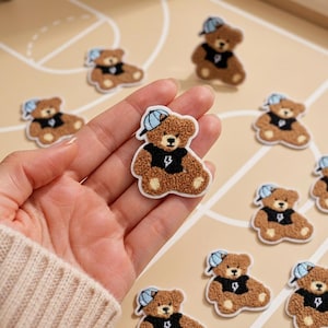 May include: A brown teddy bear patch with a textured, fuzzy surface. The bear wears a light blue baseball cap and a black shirt featuring a white lightning bolt graphic. The patch has a white border and is shown in a repeating pattern.