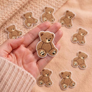 Könnte beinhalten: Eine Sammlung kleiner, brauner, flauschiger Teddybär-Aufnäher mit gestickten Details. Jeder Aufnäher zeigt einen sitzenden Bären mit strukturierter Oberfläche, dunkelbraunen Augen, einer schwarzen Nase und Pfotenabdrücken.
