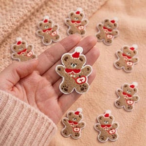 May include: A collection of small, tan, fuzzy bear-shaped patches with embroidered details. Each bear wears a red Santa hat and a red scarf, and holds a red apple. The patches have a white border and are arranged on a textured, light-colored fabric.