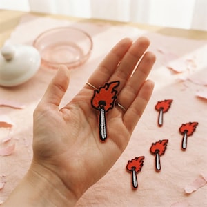 Può includere: Una toppa ricamata a forma di fiammifero acceso con una fiamma rosso brillante e un bastoncino bianco e nero. La toppa presenta un bordo nero spesso e cuciture dettagliate, che mostrano un design vivace di un fiammifero.