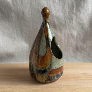 May include: A teardrop-shaped ceramic vessel with a narrow neck and a rounded top. The exterior features a mottled glaze in shades of sage green, deep brown, and black, with a smooth, glossy finish and a large, open side aperture.