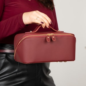 May include: A rectangular burgundy cosmetic bag with a textured surface, featuring a top handle and a gold-toned double zipper closure. The bag has a structured design with rounded corners and matching burgundy zipper pulls for easy access to the interior.