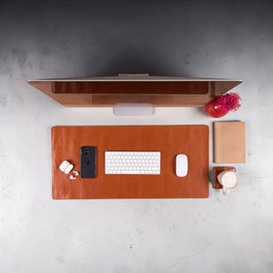 Op de afbeelding: Een rechthoekige bureaulegger van bruin leer op een oppervlak, voorzien van een wit draadloos toetsenbord, een witte computermuis, een zwarte smartphone, witte draadloze oordopjes in een hoesje, een bruin notitieboek en een witte mok met roze bloemen.